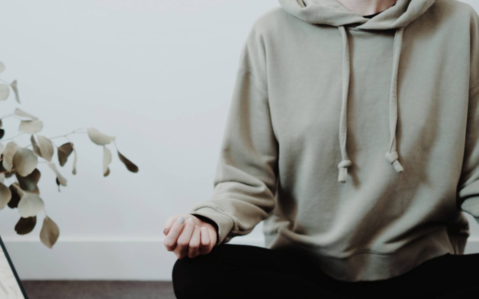 Eine Person sitzt im Schneidersitz und hält eine Hand locker geöffnet auf dem Knie. Der Bildausschnitt zeigt den Oberkörper in einem hellen Hoodie vor ruhigem Hintergrund.