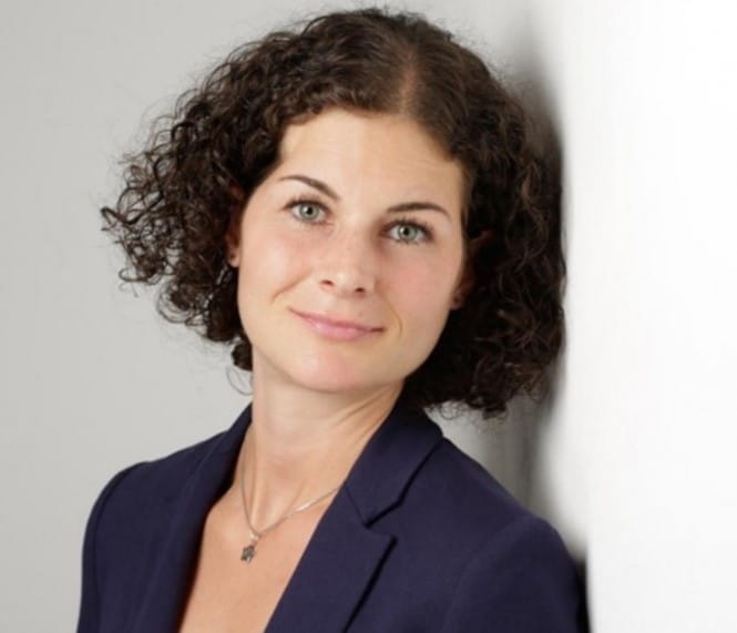 Porträt einer Frau mit lockigem dunklem Haar und dunkelblauem Blazer vor hellem Hintergrund.
