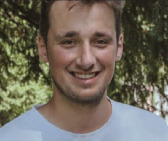 Porträt eines lächelnden Mannes mit hellem T-Shirt vor grünem Hintergrund im Freien.