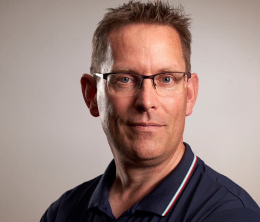 Porträt eines Mannes mit Brille und dunkelblauem Poloshirt vor hellem Hintergrund.