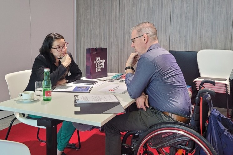 Zwei Personen sitzen konzentriert an einem Tisch und unterhalten sich. Eine der Personen nutzt einen Rollstuhl.