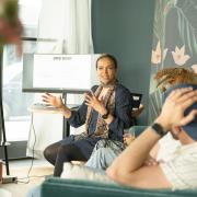 Auf dem Bild ist mittig Bisseh Akamé abgebildet; sie ist Director Diversity, Inclusion & Belonging bei UMG. Sie sitzt vor Mitarbeitenden, die ihr aufmerksam zuhören, während ihre Hände in einer erklärenden Geste erhoben sind. Auf dem Hintergrund ist ein Bildschirm, auf der Text "UMG Wert" zu lesen ist.