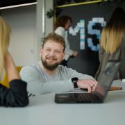 Zwei Personen unterhalten sich an einem Tisch sitzend. Im Vordergrund ein Laptop, im Hintergrund zwei weitere Personen an einer Wand mit einem MSD-Schriftzug.